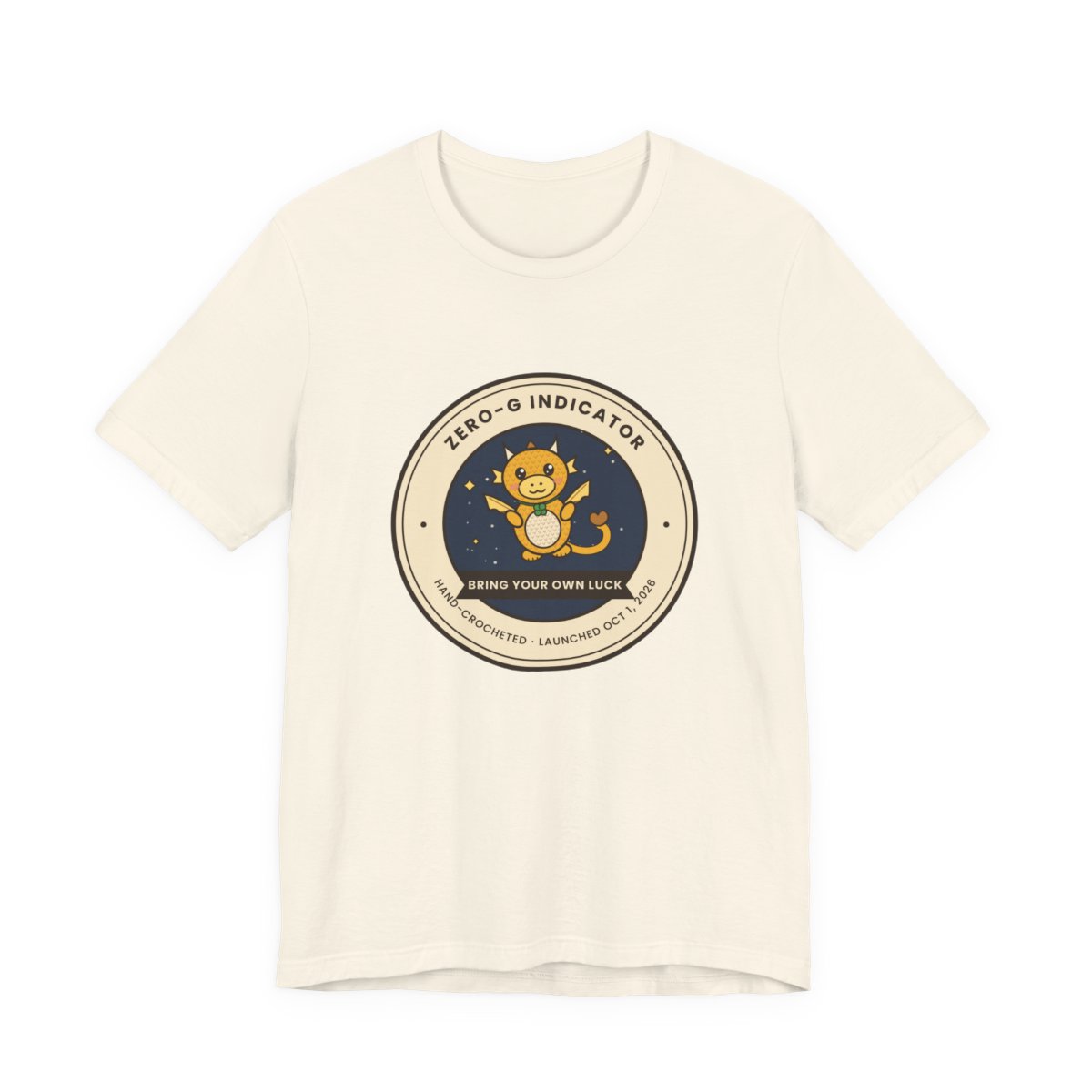 Natural-color t-shirt with a round badge: a golden crocheted dragon among stars, ringed by ZERO-G INDICATOR, with a ribbon reading BRING YOUR OWN LUCK