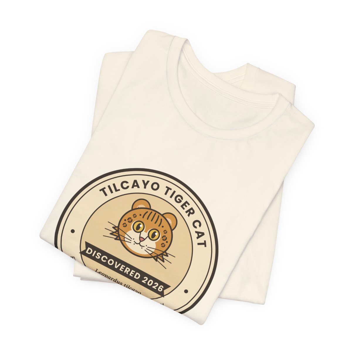 Folded natural-color t-shirt showing the round Tilcayo Tiger Cat badge with an illustrated spotted cat and a Discovered 2026 ribbon